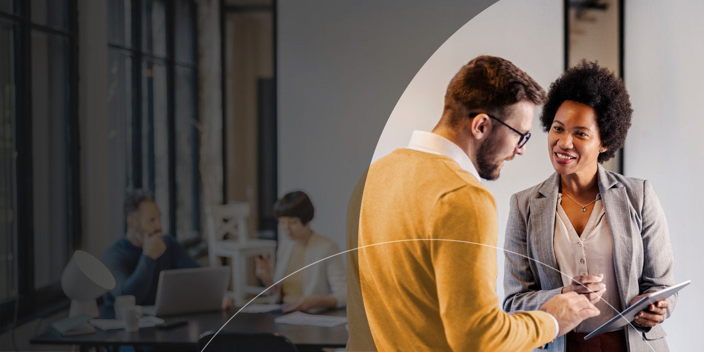 Two people engage in a conversation in an office environment, while holding a tablet.