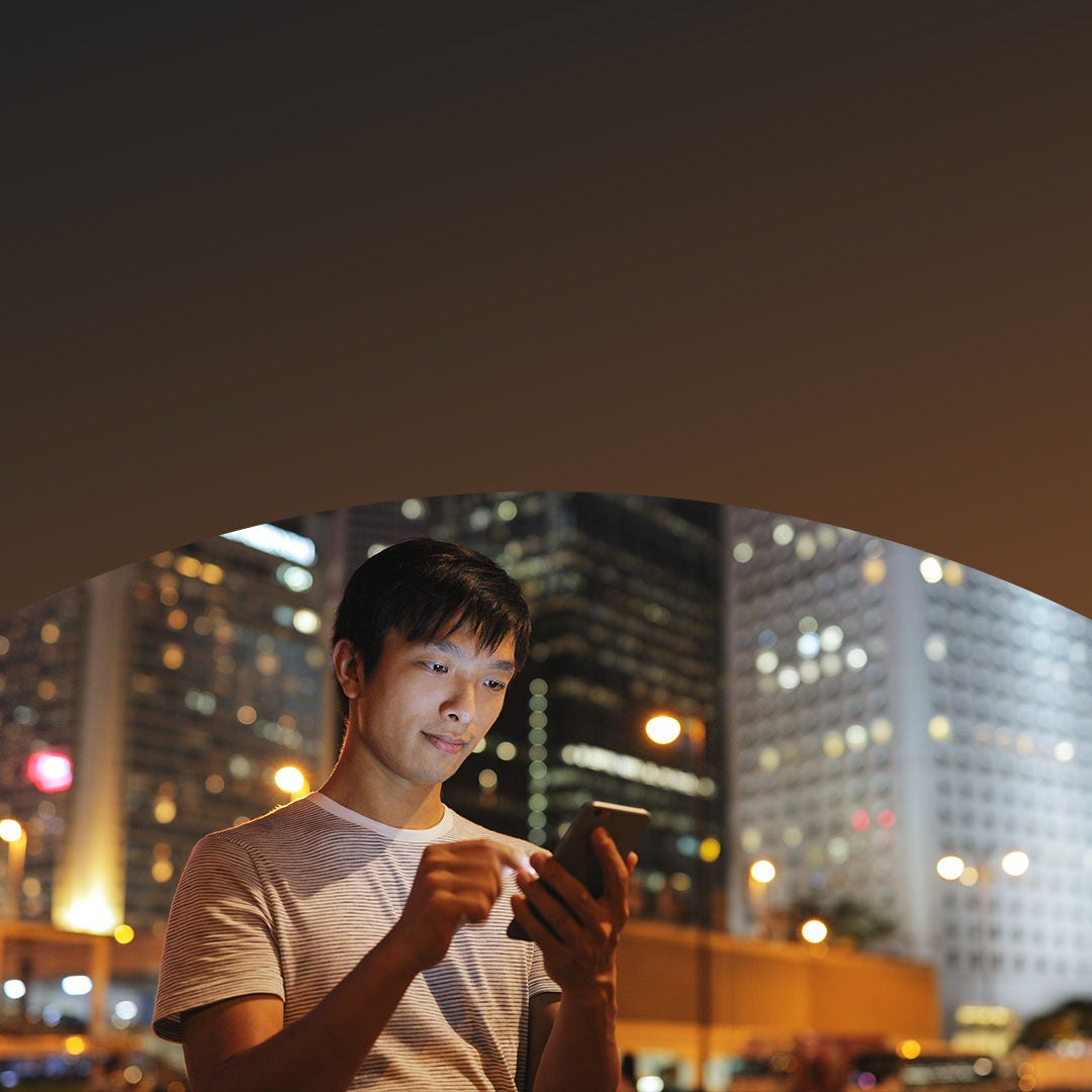 A man stands in the city, focused on his phone while surrounded by urban buildings and bustling activity.
