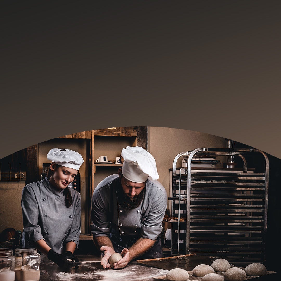 Two chefs in a bakery kitchen preparing and inspecting dough balls on the counter.