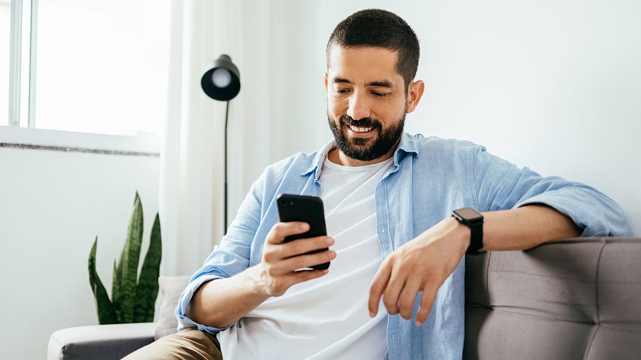 Man sitting on couch at home, using laptop and cell.
