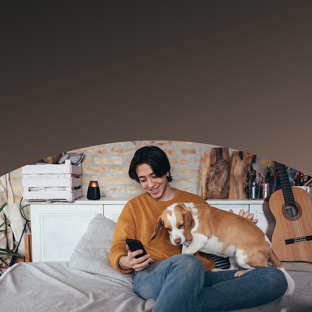 A man and his dog sit on a bed, with the man holding a cell phone in his hand, both looking relaxed.